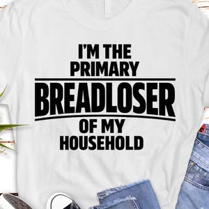 Peut inclure: T-shirt blanc avec l'inscription en noir gras : "I'M THE PRIMARY BREADLOSER OF MY HOUSEHOLD". Le texte est centré et disposé sur trois lignes, avec une ligne séparant les deux premières.
