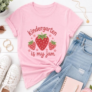 Peut inclure: Un t-shirt rose clair à manches courtes orné d'un motif de trois fraises et du texte rouge Kindergarten is my jam. La fraise centrale est grande, entourée de deux plus petites, le tout décoré de petites étoiles et d'un motif à carreaux.