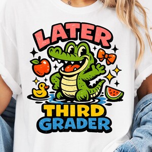 Könnte beinhalten: Weißes T-Shirt mit einem Cartoon-Alligator-Design und dem Text "LATER THIRD GRADER". Der Alligator ist grün mit gelbem Bauch, und das Design umfasst einen Apfel, eine Ente, eine Fliege und eine Wassermelone.