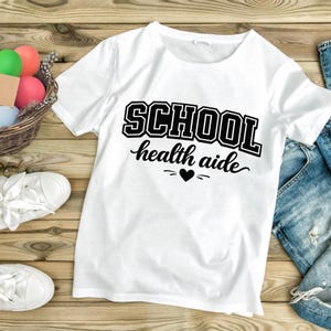 Puede incluir: Camiseta blanca con el texto negro "SCHOOL health aide". La palabra "SCHOOL" está en negrita, con un estilo universitario, y "health aide" está en cursiva. Un corazón negro está entre las dos palabras. La camiseta se muestra con vaqueros azules y zapatillas blancas.