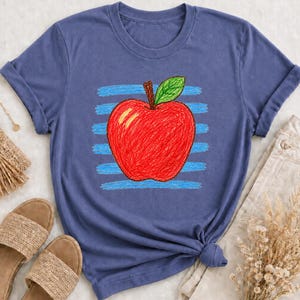 Peut inclure: Un t-shirt bleu à manches courtes arborant un motif central représentant une pomme rouge avec une feuille verte. Le dessin de la pomme présente un aspect texturé rappelant le crayon, superposé à des rayures horizontales bleu clair.
