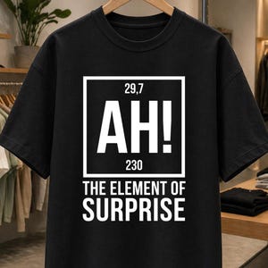 Peut inclure: T-shirt noir avec un motif blanc. Le motif présente un carré avec les chiffres "29,7" et "230", et le mot "AH!" en grandes lettres. En dessous du carré, le texte indique "THE ELEMENT OF SURPRISE".