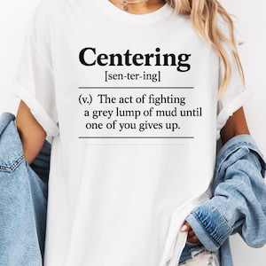May include: White t-shirt with the word "Centering" in bold black font, followed by the phonetic pronunciation. Below, a definition reads, "(v.) The act of fighting a grey lump of mud until one of you gives up."