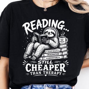 Peut inclure: T-shirt noir avec un motif blanc représentant un paresseux lisant un livre, avec le texte "READING... STILL CHEAPER THAN THERAPY." Le paresseux est assis sur une pile de livres. Une tasse à café avec le texte "PAGES OVER PROBLEMS" est à côté du paresseux.