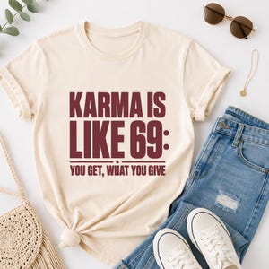 Peut inclure: Un t-shirt à manches courtes de couleur crème avec un texte rouge foncé qui indique : KARMA IS LIKE 69: YOU GET, WHAT YOU GIVE. Le t-shirt possède un col rond et est présenté avec un jean bleu, des baskets blanches, des lunettes et un collier.