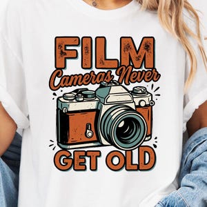 Op de afbeelding: Wit T-shirt met een vintage camera-afbeelding en de tekst "FILM Cameras Never GET OLD" in bruin en oranje. Het ontwerp heeft een retro esthetiek, voor fotografie liefhebbers.