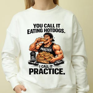 Op de afbeelding: Witte sweatshirt met een afbeelding van een gespierde man die een hotdog eet, met de tekst "YOU CALL IT EATING HOTDOGS. I CALL IT PRACTICE." De afbeelding bevat een dienblad met hotdogs, dumbbells en de woorden "EAT TRAIN REPEAT."