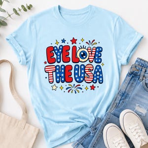 May include: A light blue short-sleeve t-shirt featuring a graphic print with the text EYE LOVE THE USA. The design incorporates a cartoon eyeball, red and white stripes, and blue stars, accented by small red, yellow, and blue star and firework icons.