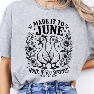 Peut inclure: Un t-shirt gris chiné avec une impression graphique noire représentant deux canards entourés d'éléments floraux. Le texte indique "MADE IT TO JUNE" en haut et "HONK IF YOU SURVIVED" en bas, avec de petites étoiles et des ornements.