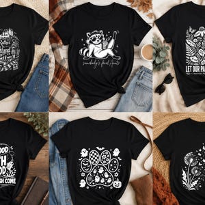 Peut inclure: Une collection de six t-shirts noirs avec divers motifs graphiques blancs. Les designs incluent un paysage d'Écosse avec du texte, un raton laveur avec du texte, un graphique floral de parcs nationaux, un thème de pêche, une patte avec des fantômes et des pissenlits.
