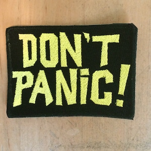 Peut inclure: Un patch en tissu noir avec du texte brodé jaune qui dit "DON'T PANIC!"