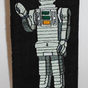 May include: Embroidered patch featuring a retro robot design in shades of gray, white, and orange on a black background. The robot has a blocky design with segmented limbs and a rectangular head. The robot is waving.