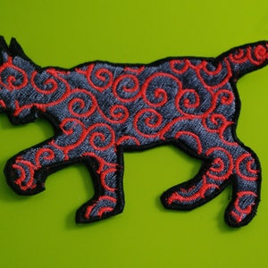 Può includere: Toppa ricamata a forma di gatto, con sfondo grigio scuro e motivi a spirale rossi. Il gatto è in movimento, con contorni e zampe neri. La toppa è su uno sfondo verde brillante.