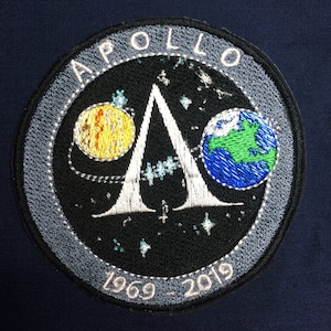 May include: A grey and white embroidered patch with the text "APOLLO" in a circle around a white letter "A". The "A" is surrounded by a black background with a yellow moon, a blue and green earth, and white stars. The text "1969-2019" is at the bottom of the patch.