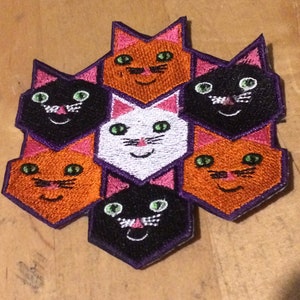 May include: A hexagonal patch with six embroidered cat faces. The cats are orange, black, and white. The cats have green eyes and pink noses. The patch is surrounded by a purple border.