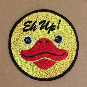 May include: Embroidered patch featuring a yellow duck face with a red beak and black eyes. The phrase "Eh Up!" is stitched in black above the duck's face. The patch has a black border and is circular in shape.
