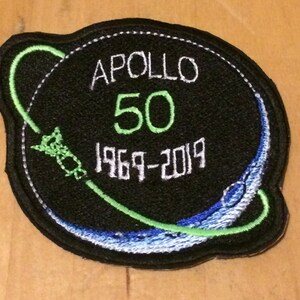 May include: A black embroidered patch with a green and white design. The patch features a stylized image of the Earth with a green orbit around it. The text "APOLLO 50 1969-2019" is embroidered in white on the patch.