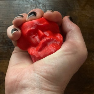 Könnte beinhalten: Ein leuchtend rotes, schädelartiges Objekt wird in einer Hand gehalten. Der Schädel hat detaillierte Gesichtszüge und eine strukturierte Oberfläche. Die Hand hat schwarzen Nagellack. Der Hintergrund ist eine dunkle Holzoberfläche.