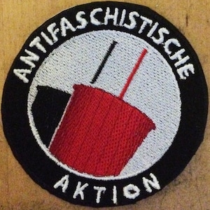 May include: Black and white embroidered patch with the text "Antifa Schistische Aktion" in white. The patch features a red cup with a black straw and a white straw.