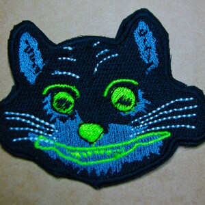 May include: Black cat face embroidered patch with green eyes and a blue and green grin. The patch has white stitching for whiskers.