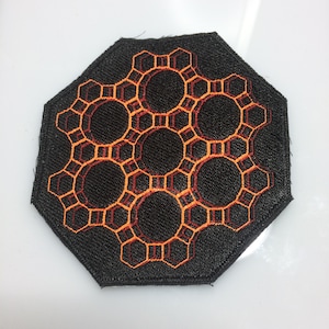 Può includere: Una toppa ottagonale nera con un disegno geometrico ricamato arancione e rosso. Il motivo ricorda un favo o una struttura molecolare.