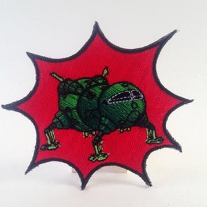 May include: A green embroidered patch of a cartoon spaceship with three legs, set against a red background with a black outline. The patch is shaped like a starburst.