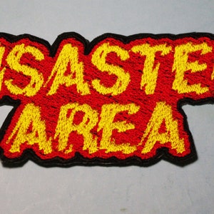 以下が含まれることがあります： 赤い背景に黄色い刺繍で「Disaster Area」と書いてあります。