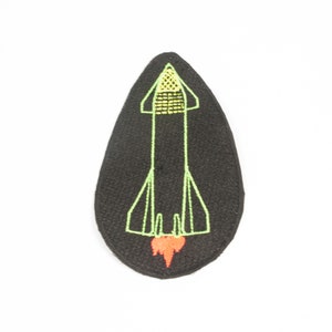 May include: A black felt patch with an embroidered green rocket ship with a yellow top and an orange flame.