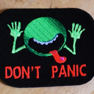 Puede incluir: Un parche bordado negro con un extraterrestre verde con una lengua roja y el texto "DON'T PANIC" en rojo.