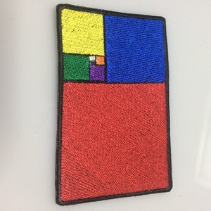 Può includere: Una toppa ricamata con un design geometrico colorato ispirato al rapporto aureo. La toppa è composta da quadrati in tonalità di giallo, blu, verde, arancione, viola e rosso.