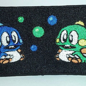 May include: Embroidered patch featuring two cartoon dinosaurs, one blue and one green, with white bellies and yellow accents. They are surrounded by green and blue bubbles on a black background.