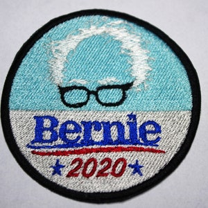 May include: A round, black-bordered, embroidered patch with a blue background. The patch features a white outline of a person's hair, black-rimmed glasses, and the text "Bernie 2020" in blue, red, and white.