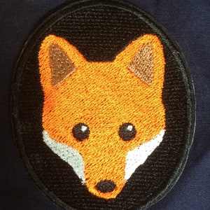 Pode incluir: Patch bordado de cabeça de raposa laranja com contorno preto e detalhes brancos sobre fundo azul escuro.