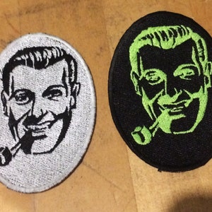 Puede incluir: Dos parches bordados con un hombre con una pipa. Un parche es blanco con bordado negro, el otro es negro con bordado verde.