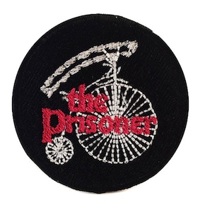 May include: A black embroidered patch with a white bicycle and the text "The Prisoner" in red.