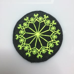 May include: A black circular patch with a green embroidered design. The design features a circular pattern with a starburst in the center.