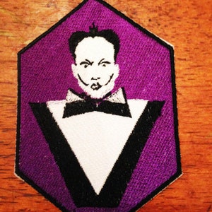 Puede incluir: Un parche hexagonal morado con un diseño de esmoquin blanco y negro. El esmoquin tiene una pajarita blanca y una camisa blanca con un contorno negro. Una ilustración en blanco y negro de la cara de una persona está en el centro del parche.