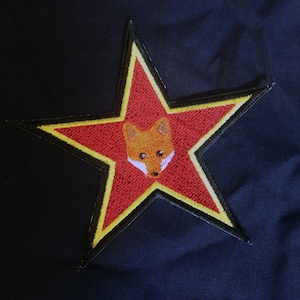 Könnte beinhalten: Ein roter Stern mit gelbem Rand und einem Fuchsgesicht in der Mitte. Der Stern ist auf einem dunkelblauen Stoff gestickt.