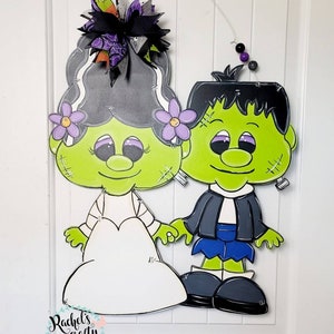 Può includere: Un appendino per porta di Halloween verde e nero con una sposa e uno sposo vestiti da mostro di Frankenstein. La sposa indossa un abito bianco e un fiore viola nei capelli. Lo sposo indossa un abito nero e una cravatta blu. L'appendino per porta è decorato con un fiocco nero e un fiore viola e nero.