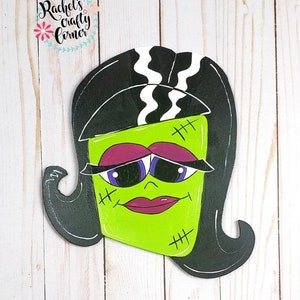 Può includere: Un ritaglio di legno verde e nero di un mostro di Frankenstein con capelli neri, linee a zig-zag bianche e punti di sutura sul viso. Il mostro ha occhi viola e una bocca rosa.