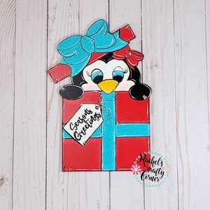 A penguin wearing a blue bow and red bow peeking out of a red gift box with a blue ribbon and a tag that says 'Seasons Greetings'. The gift box is on a white wood background.