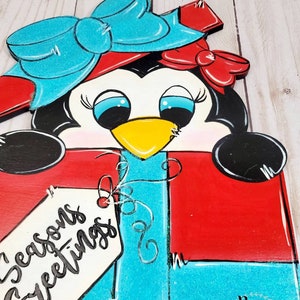 A penguin wearing a red and blue bow and a red hat is holding a red gift box with a blue ribbon and a white tag that says 'Seasons Greetings'. The gift box is decorated with white stitching and a white swirl design.