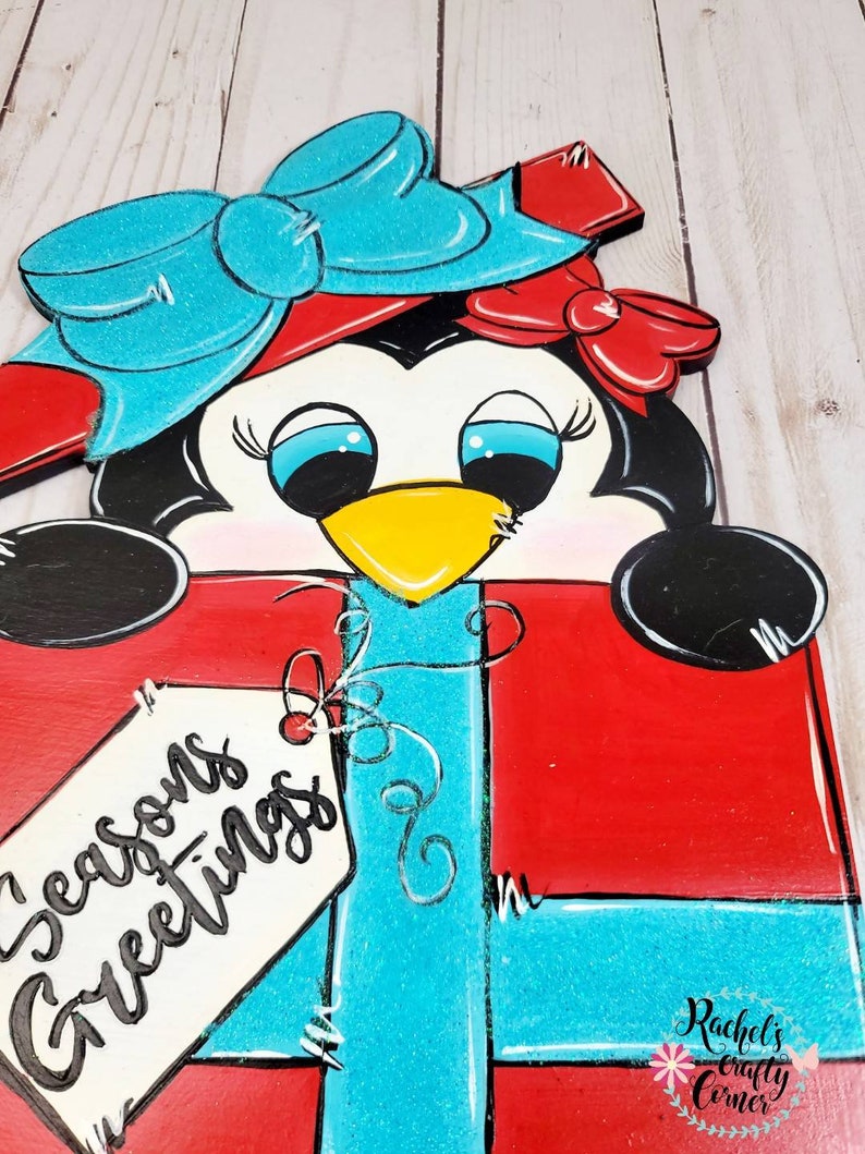 A penguin wearing a red and blue bow and a red hat is holding a red gift box with a blue ribbon and a white tag that says 'Seasons Greetings'. The gift box is decorated with white stitching and a white swirl design.