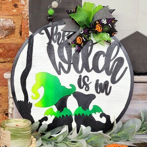 Puede incluir: Un letrero redondo de madera blanca con el texto "The Witch is in" en letras negras. Dos siluetas de brujas negras con sombreros y alas verdes están en el letrero. Un lazo verde y negro con un estampado de Halloween está unido a la parte superior del letrero.