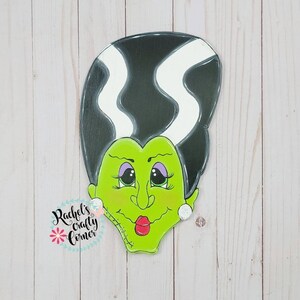 Può includere: Un ritaglio di legno dipinto di verde e nero di una donna Frankenstein con cuciture bianche sul viso. Il ritaglio ha occhi grandi con palpebre viola e una bocca rossa. Il ritaglio è su uno sfondo di legno bianco.