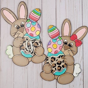 Könnte beinhalten: Zwei braune Hasen-Türhänger mit bemalten Details. Jeder Hase hält ein großes, buntes Osterei mit Leopardenmuster. Ein Hase hat eine rote Schleife.