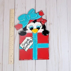 A red and blue penguin wearing a blue bow and red bow peeking out of a red gift box with a blue ribbon. The gift box has a white tag that says 'Seasons Greetings'.