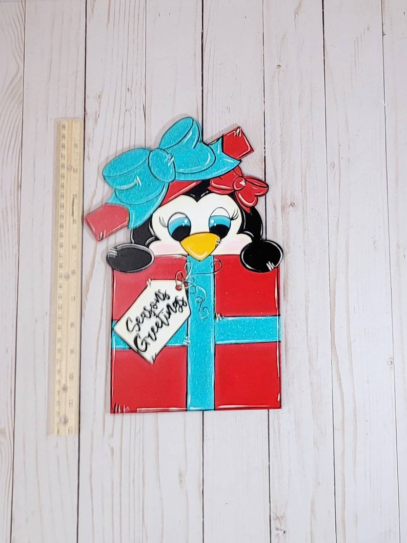 A red and blue penguin wearing a blue bow and red bow peeking out of a red gift box with a blue ribbon. The gift box has a white tag that says 'Seasons Greetings'.