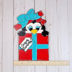 A penguin wearing a blue bow and red ribbon peeking out of a red and blue gift box. The gift box has a white tag that says 'Seasons Greetings'.