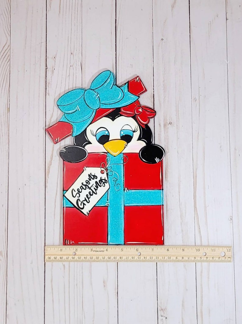 A penguin wearing a blue bow and red ribbon peeking out of a red and blue gift box. The gift box has a white tag that says 'Seasons Greetings'.